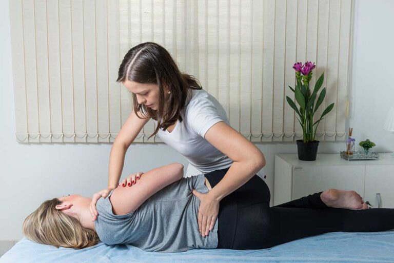 A person sits on a bench, massaging their foot while another person checks posture and back alignment outdoors.